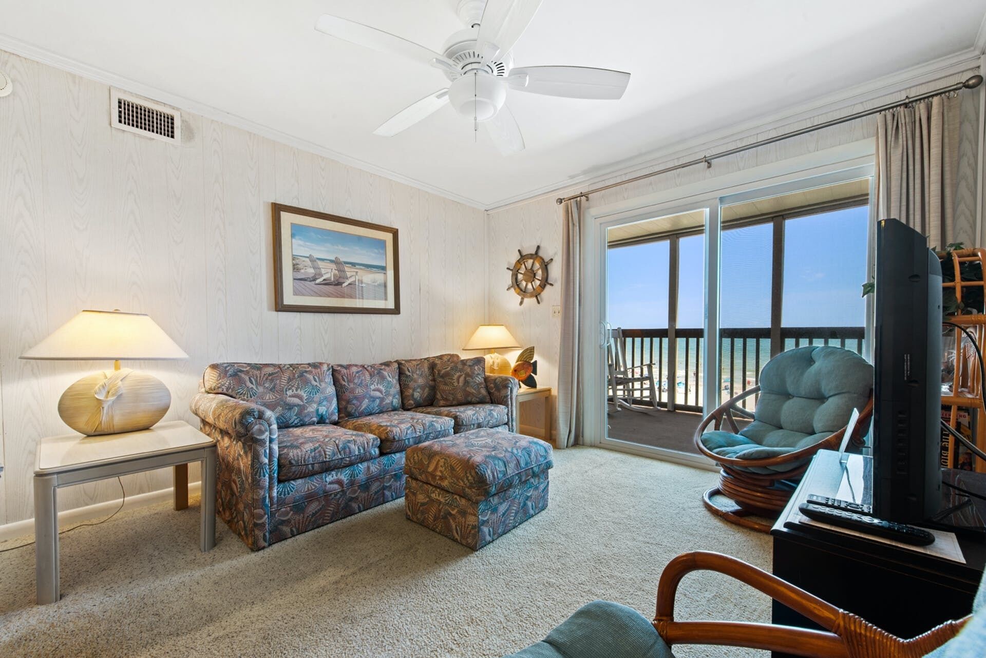A living room with a sofa, two side tables with lamps, a television, and a view of the beach through large windows.