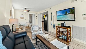 A living room with a black sofa, a dining table with chairs, a TV on the wall, and a bookshelf. - 2-bedroom Corner Unit with private balcony | Carolina Reef - 301 (North Myrtle Beach)