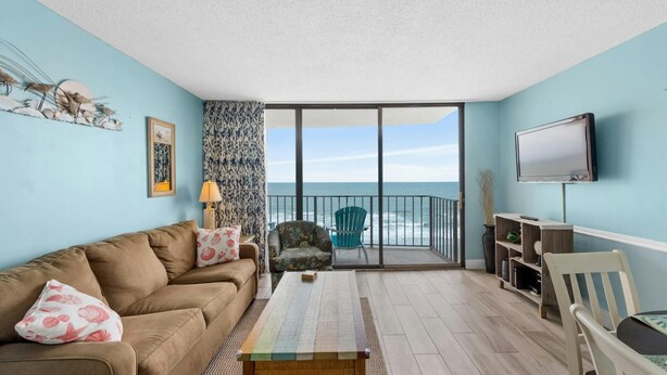A living room with a sofa, a table, a TV, and a view of the ocean. - Updated 1-Bedroom Oceanfront Condo| Carolina Reef - 704 (North Myrtle Beach)