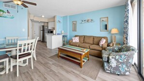A living room with a sofa, a coffee table, and a patterned armchair. There is a dining area with a table and chairs. The room has light blue walls and a ceiling fan. - Updated 1-Bedroom Oceanfront Condo| Carolina Reef - 704 (North Myrtle Beach)