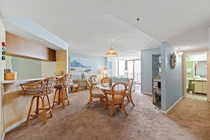 A dining area with wicker chairs and a round table, a kitchen with a view, and a living area with a large window. - Sea Lovers Delight! 3-bedroom, 2-bathroom, oceanfront condo | Sea Castle - 7B (North Myrtle Beach)