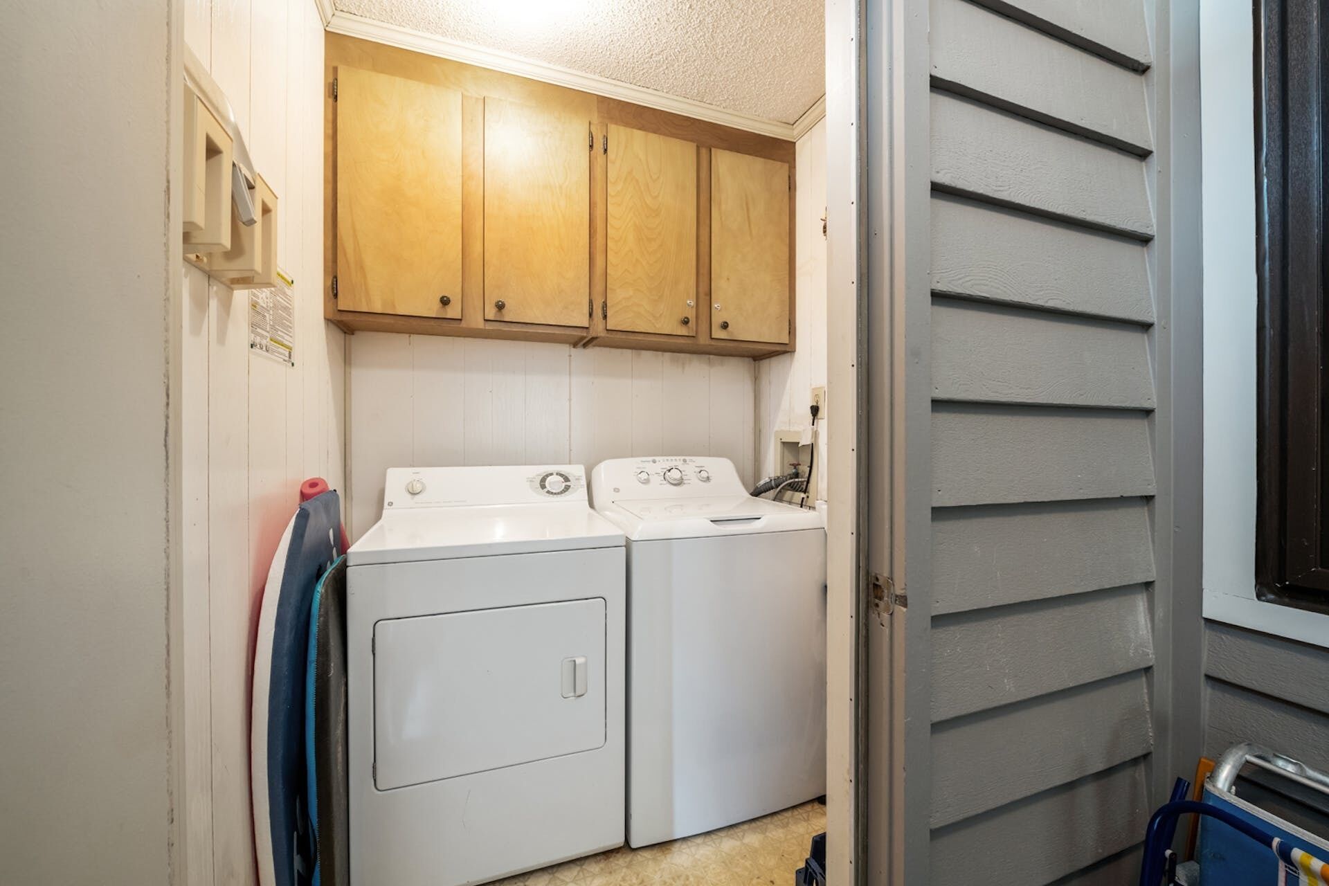 A laundry room with two white washers and dryers, wooden cabinets above, and a window with blinds.