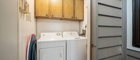 A laundry room with two white washers and dryers, wooden cabinets above, and a window with blinds.