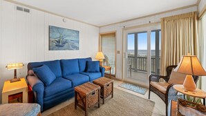 A living room with a blue sofa, two side tables with lamps, a wicker coffee table, a painting of boats, and a balcony with curtains. - 3-bedrom, 2.5 bath, 1st floor oceanfront condo | Dunescape - 101 (North Myrtle Beach)