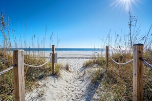 Ein sandiger Küstenweg führt zum Meer, gesäumt von Holzbefestigungen und Seilen.