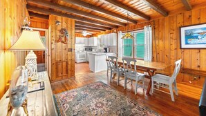 A wooden cabin interior with a dining area, kitchen, and a large window with curtains. - 3-bedroom Beach Cottage, 4th Row, Large Back Deck and screened in porch | Mermaid Cottage - 4000 (North Myrtle Beach)