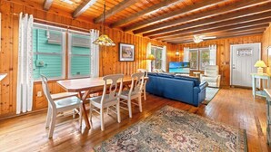 A wooden cabin interior with a dining area, sofa, and a television displaying a beach scene. - 3-bedroom Beach Cottage, 4th Row, Large Back Deck and screened in porch | Mermaid Cottage - 4000 (North Myrtle Beach)