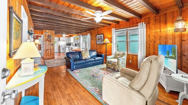 A cozy cabin interior with a wooden ceiling, a ceiling fan, a sofa, a chair, a television, and a kitchen in the background. - 3-bedroom Beach Cottage, 4th Row, Large Back Deck and screened in porch | Mermaid Cottage - 4000 (North Myrtle Beach)