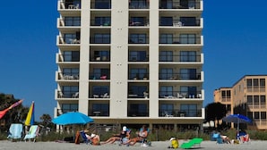 Et hotel med flere etager og altaner på hver etage, beliggende nær en strand med liggestole og parasoller.