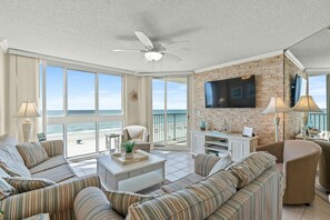 A spacious living room with a large window overlooking the beach, a ceiling fan, and a flat-screen TV mounted on a brick wall. - Luxury 2-bedroom, 2-bathroom oceanfront condo, 4th floor | Pinnacle - 404 (North Myrtle Beach)