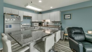 A modern kitchen with white cabinets, a granite countertop, and stainless steel appliances. There is a black leather chair and a small table with a phone. - Renovated 1-bedroom, 1-bath 3rd floor oceanfront condo | Ocean Inn - 303 (North Myrtle Beach)