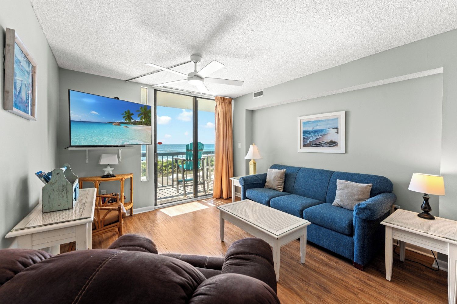 A living room with a blue sofa, a coffee table, a TV, and a balcony with a view of the ocean.