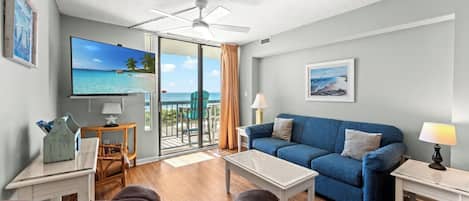 A living room with a blue sofa, a coffee table, a TV, and a balcony with a view of the ocean.
