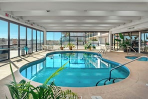 Una piscina interna con pareti di vetro trasparente, zona solarium con sedie e vista sugli edifici circostanti.