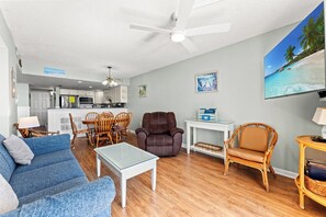 A living room with a sofa, armchair, dining table, and chairs. There is a kitchen in the background with white cabinets and a ceiling fan.