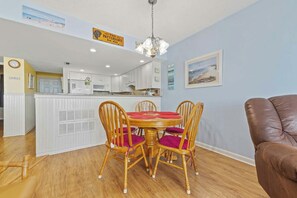 A dining area with a round table and four chairs, a kitchen in the background, and a brown leather chair to the right.