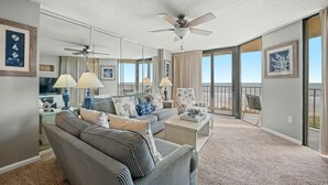 A spacious living room with a large sofa, a coffee table, and a ceiling fan. There are two lamps on the side table, and a large window with a view of the beach. - Calming paint palette and stylish design, 3-bedroom, 3-bath, sleeps 8, oceanfront condo | Summit -3D (North Myrtle Beach)