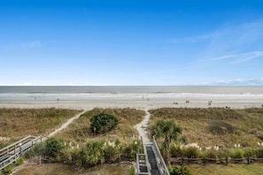 A beach with a wooden boardwalk leading to the shore, surrounded by grassy dunes and sparse vegetation.