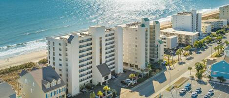 A coastal hotel with multiple buildings, a parking lot, and a beach view.