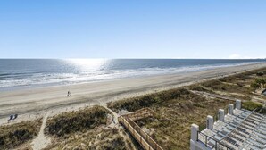 A beach with a wooden walkway leading to a sandy area, a clear sky, and the ocean in the background.