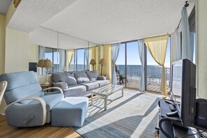 Amplia sala de estar con sofá, reposapiés y una mesa de centro. Hay un televisor y un sistema de sonido sobre un mueble bajo. La habitación tiene ventanas grandes con cortinas y vistas al océano.