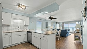 Une cuisine moderne avec des meubles blancs, un plan de travail en marbre et un coin repas agrémenté d’un canapé bleu et d’un ventilateur de plafond.