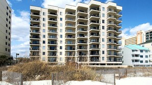Un immeuble résidentiel à plusieurs étages, doté de balcons, avec un rez-de-chaussée sablonneux et un ciel dégagé.