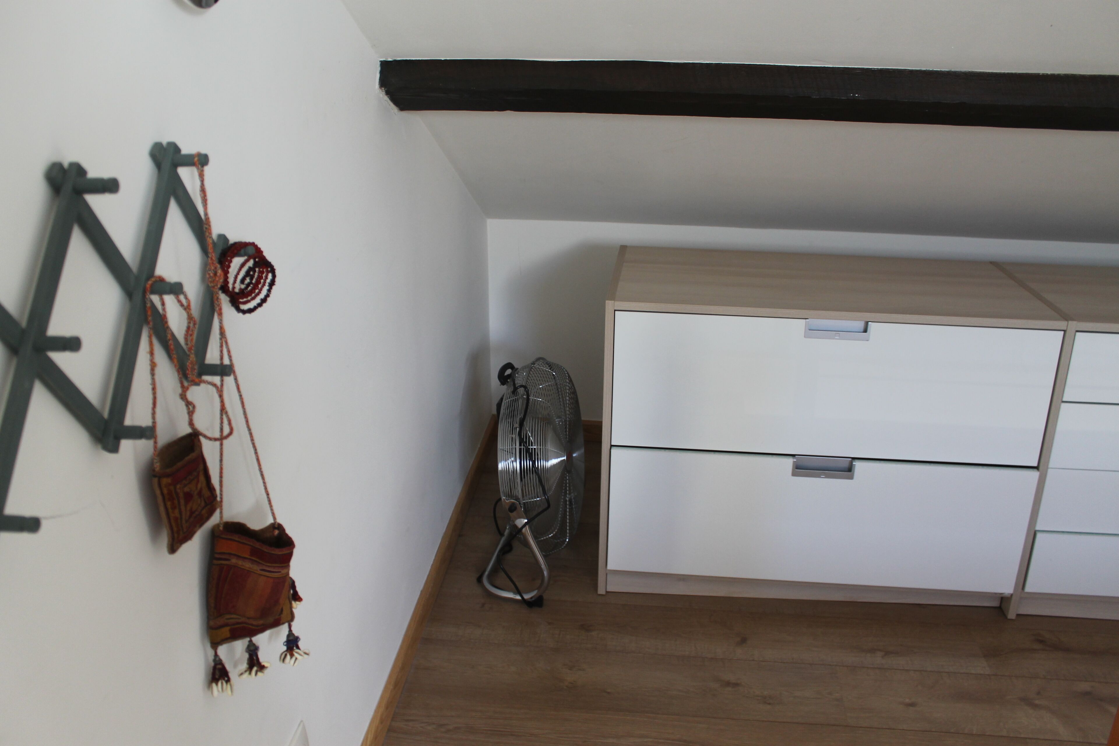 Een kamer met een houten vloer, een wit kastje, een ventilator en een wandrek met tassen.