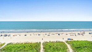 Ein weiter Sandstrand mit Menschen und Sonnenschirmen, ein klarer Himmel und ruhiges Meer.