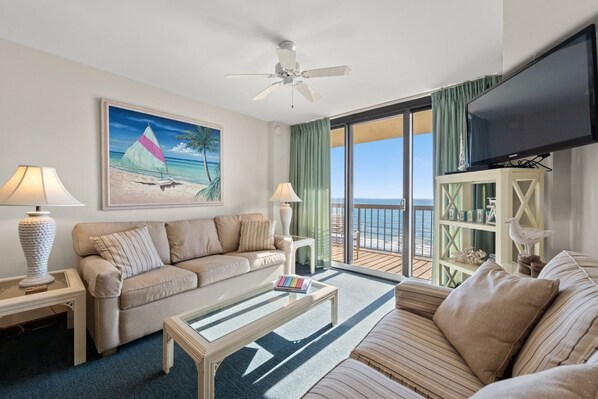 A hotel room with a sofa, a coffee table, a TV, a painting of a beach scene, and a balcony with a view of the ocean.