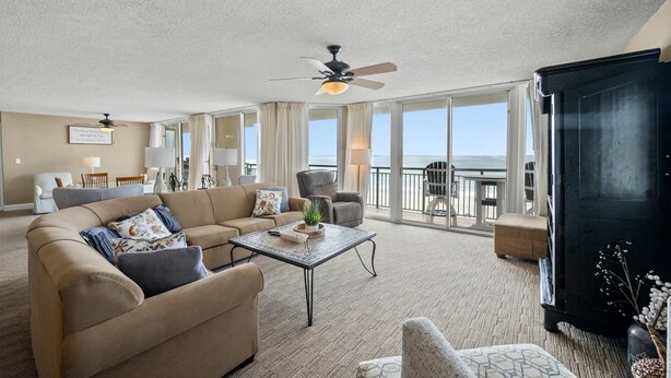 Ein geräumiges Wohnzimmer mit einem beigefarbenen Sofa, einem Couchtisch und einem Deckenventilator. Ein Balkon bietet Meerblick.