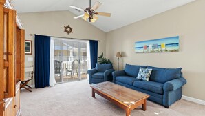 A living room with a blue sofa, a wooden coffee table, a ceiling fan, and a painting of beach chairs. - Golf Course Views, 2-bed, 2-bath Condo, Barefoot Resort | Ironwood - 834 (North Myrtle Beach)