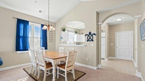 A dining area with a wooden table and white chairs, a chandelier, and a kitchen area with white cabinets and a blue wall decoration. - Golf Course Views, 2-bed, 2-bath Condo, Barefoot Resort | Ironwood - 834 (North Myrtle Beach)