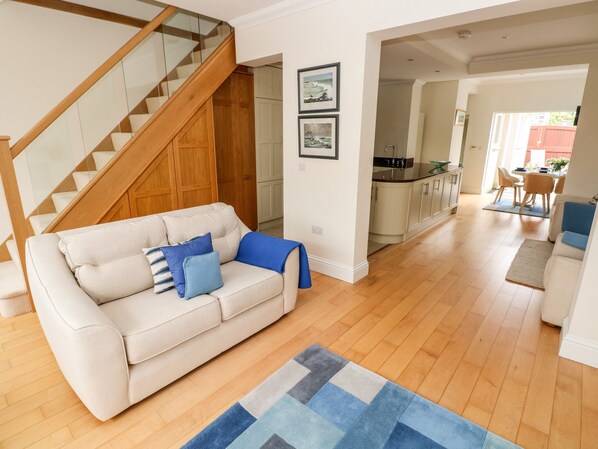 A living room with a beige sofa, a wooden staircase, a kitchen, and a dining area.