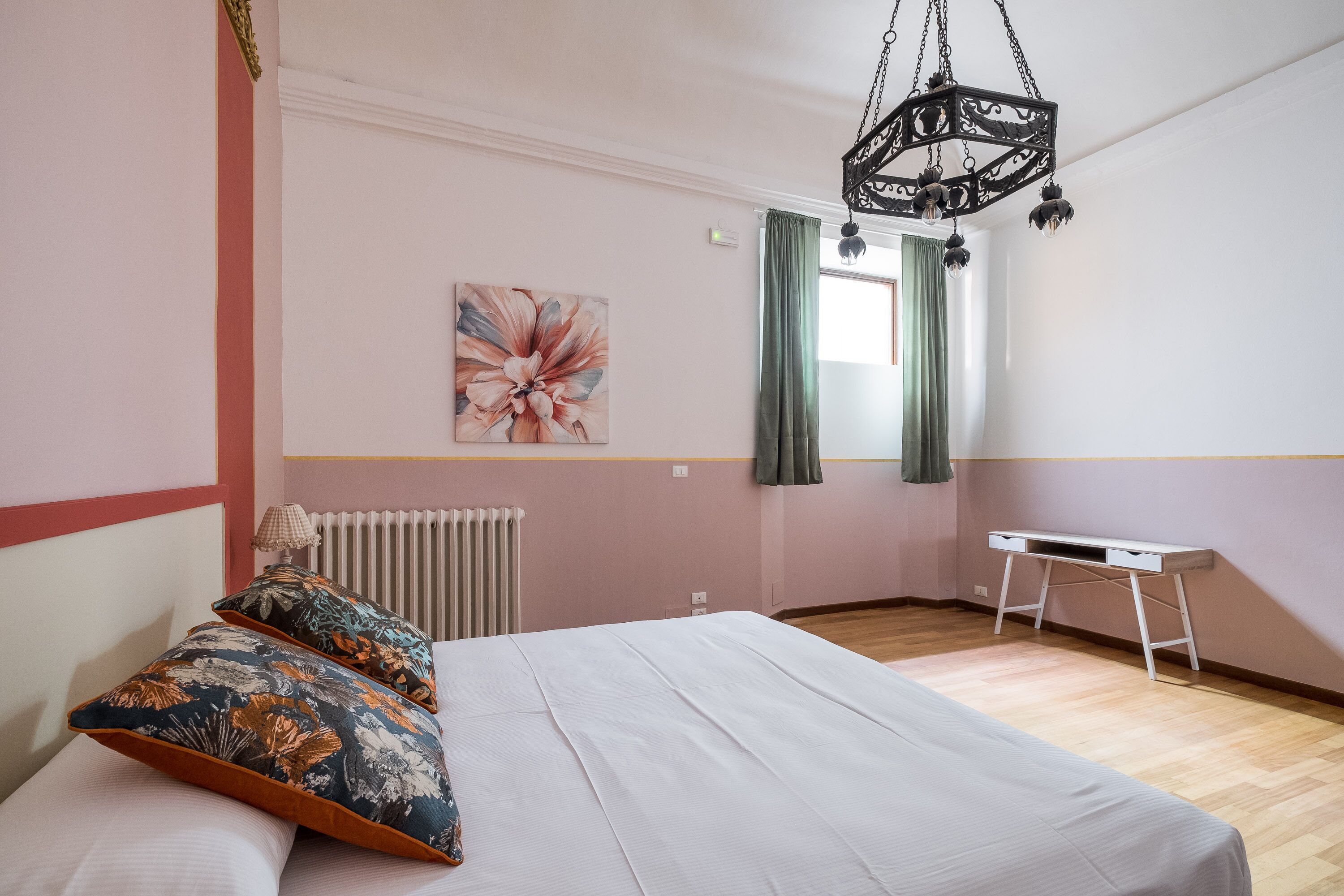 A bedroom with a bed, a desk, a radiator, a window with curtains, and a hanging light fixture.