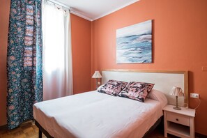 Une chambre à coucher avec un lit, une table de chevet, une lampe, un tableau et une fenêtre avec des rideaux.