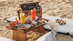 A beach picnic setup with a wooden table, a wicker basket, and a variety of food and drinks. - Melia Phuket Mai Khao (Mai Khao)