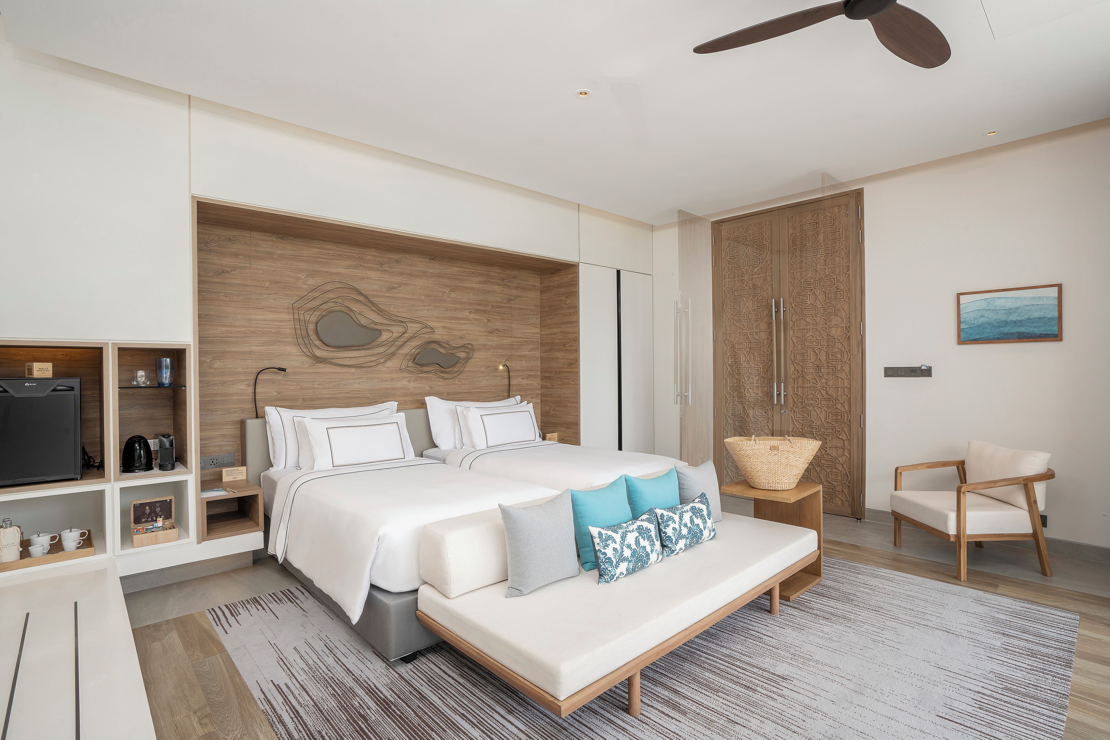 Un dormitorio moderno con una cama grande, cabecera de madera, ventilador de techo, televisor y un espacio de descanso con una silla y una mesita.