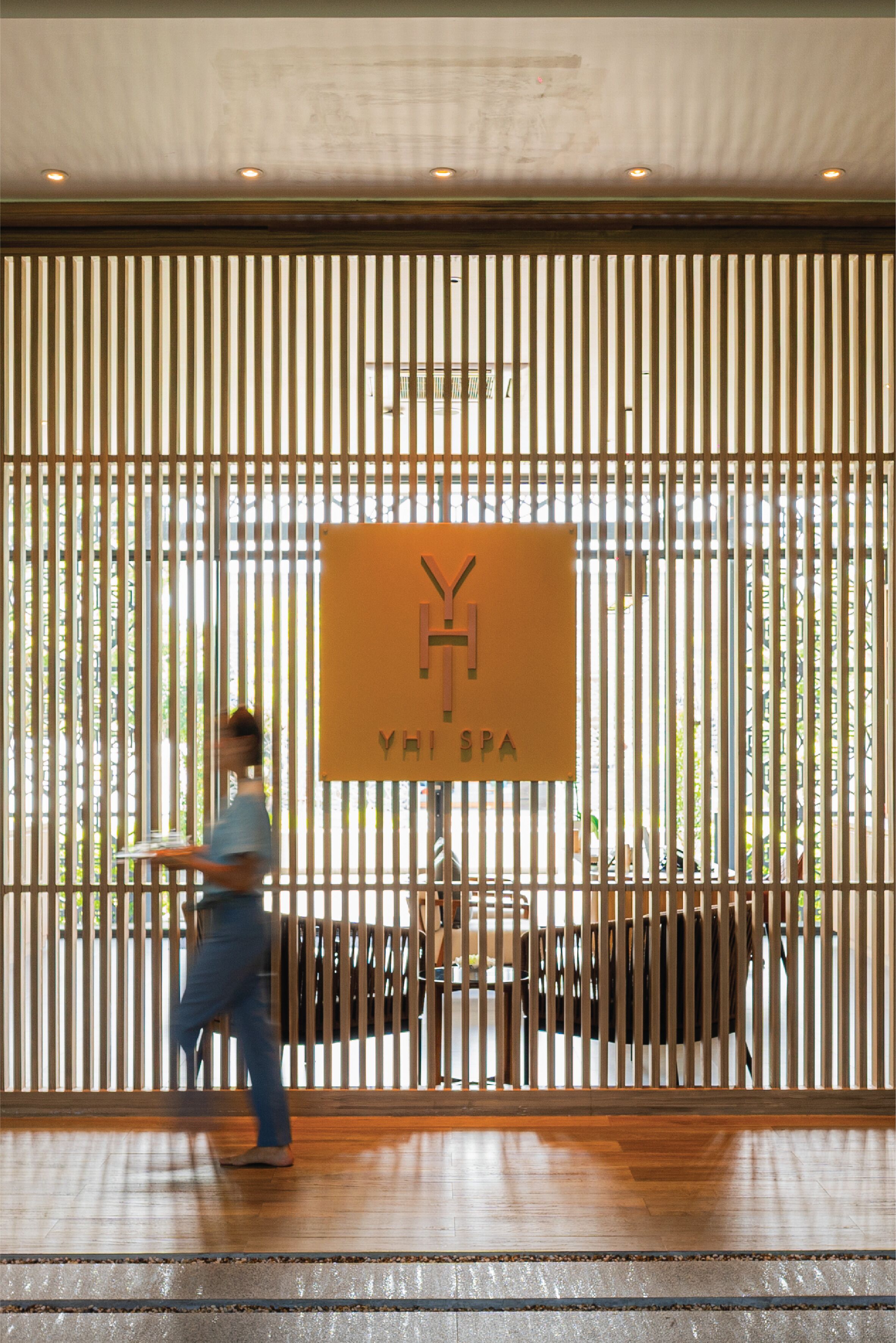 A modern interior with vertical wooden slats, a sign reading "YHI SPA", and a person carrying a tray.