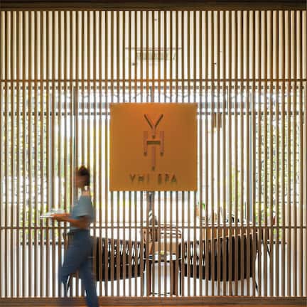 A modern interior with vertical wooden slats, a sign reading "YHI SPA", and a person carrying a tray.