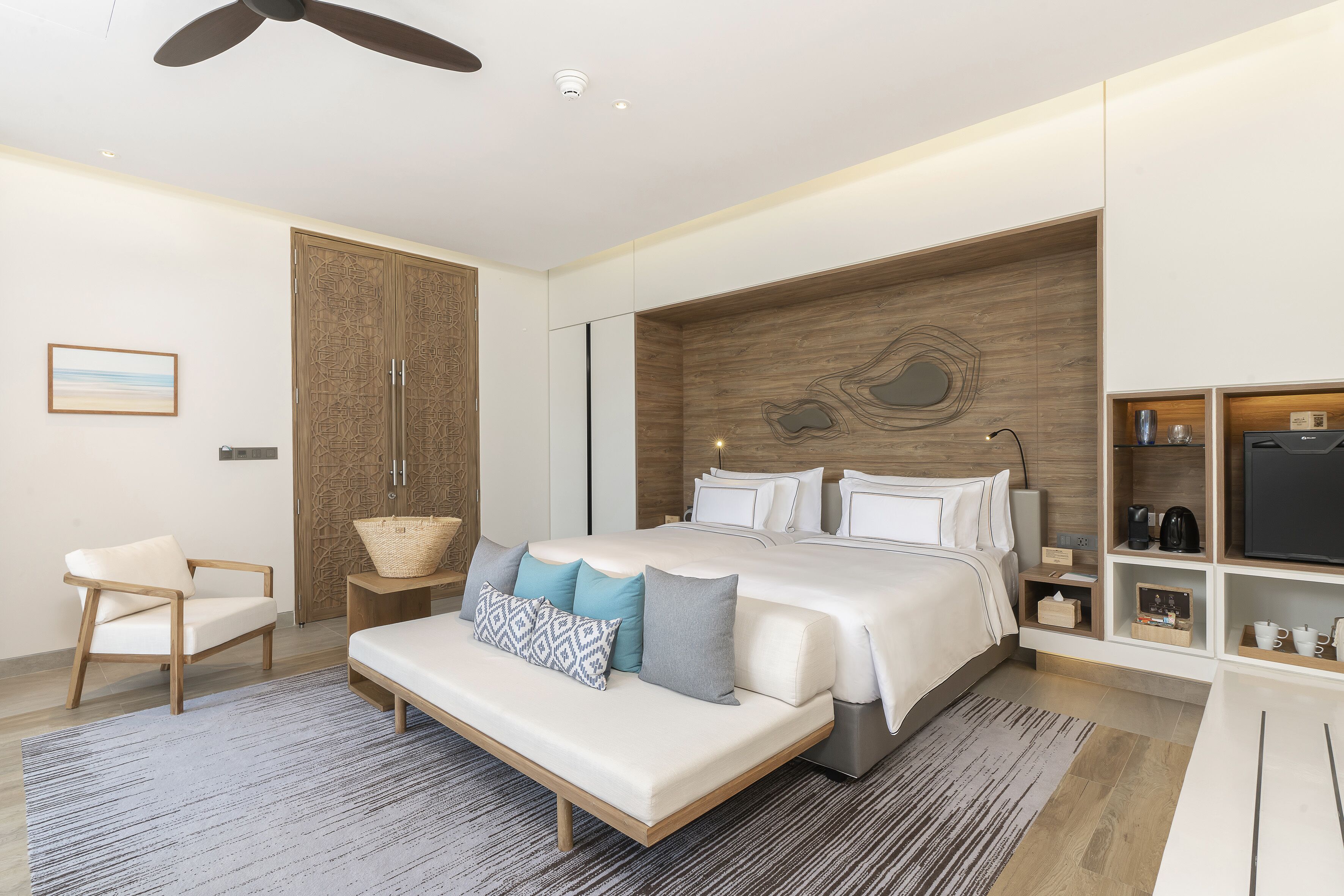A modern bedroom with a large bed, a wooden headboard, a seating area with a bench, and a TV on a shelf.