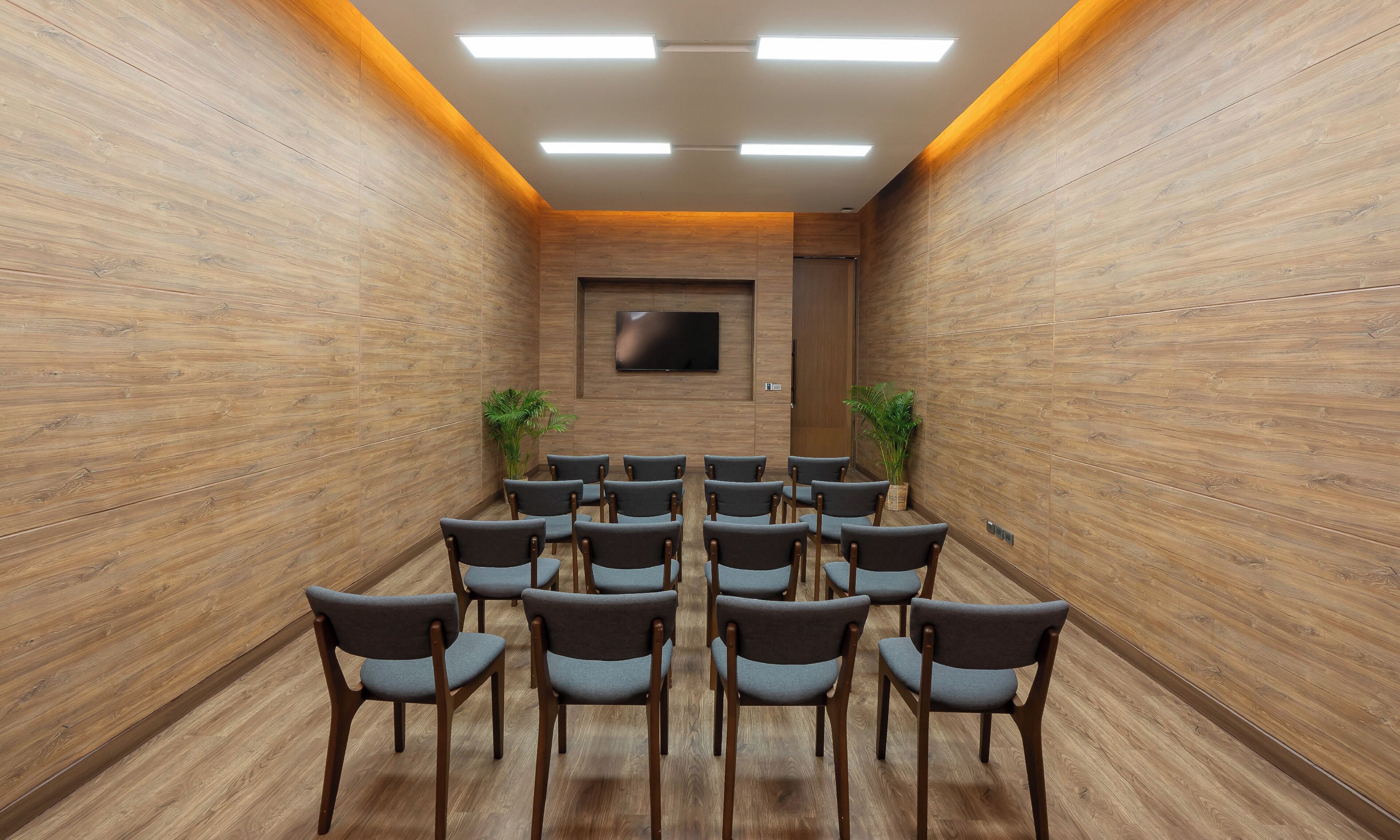 A conference room with wooden walls, a flat-screen TV, and rows of grey chairs.