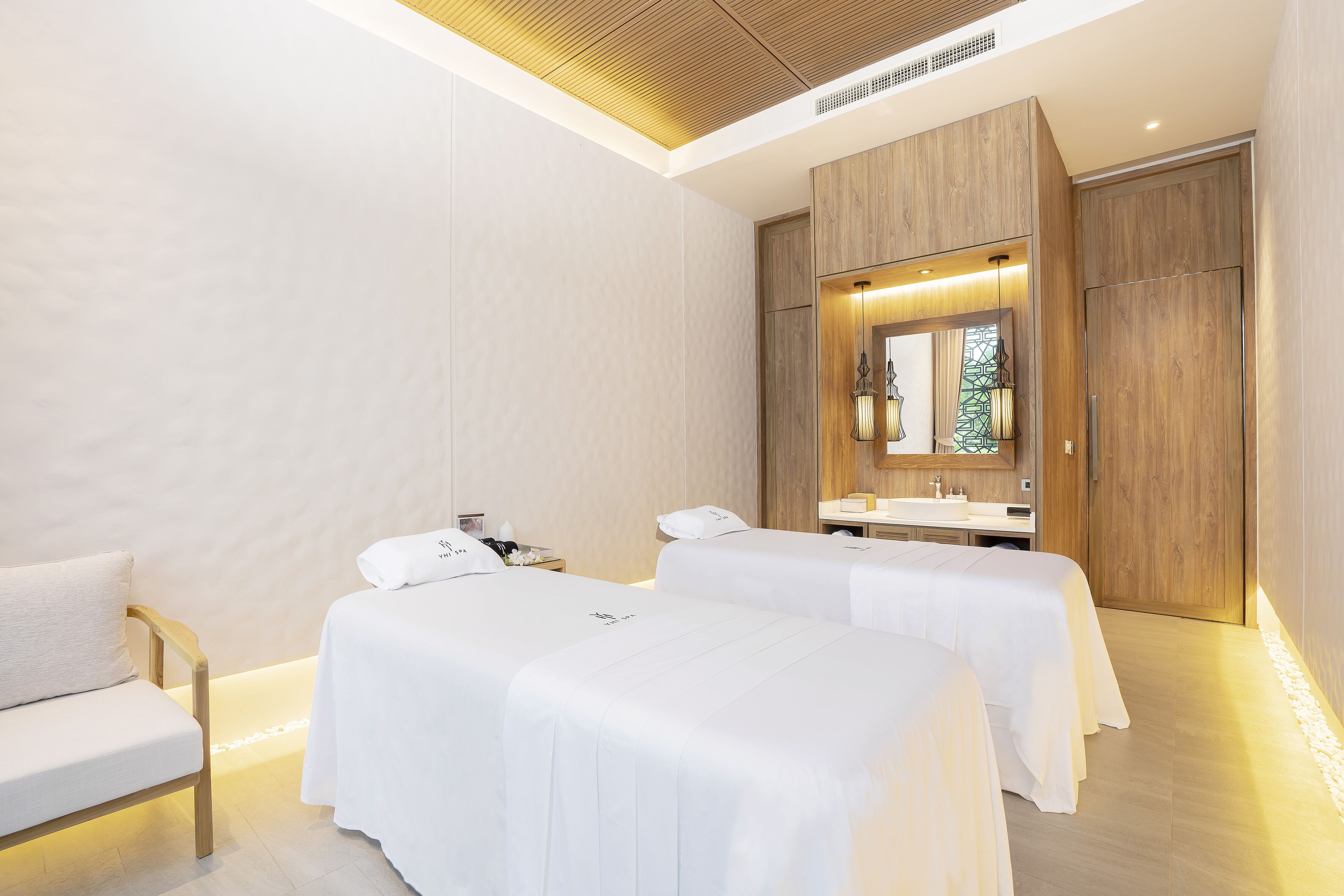 A modern hotel room with two beds, a sofa, a vanity with a mirror, and a wooden ceiling.
