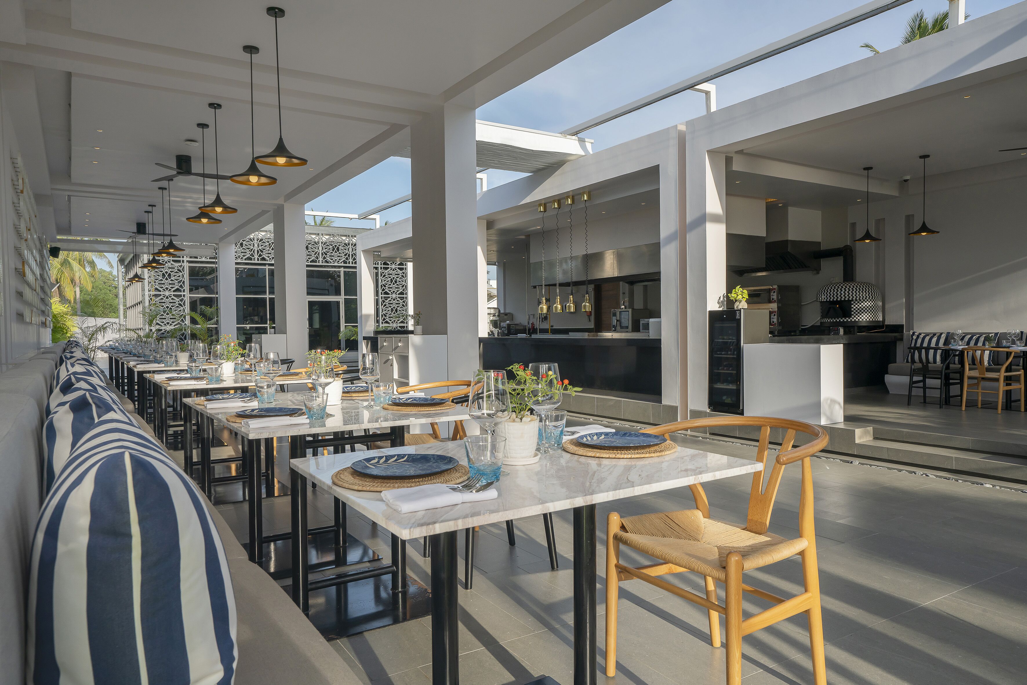 A modern outdoor dining area with multiple tables and chairs, a kitchen area, and a seating area with a striped cushion.