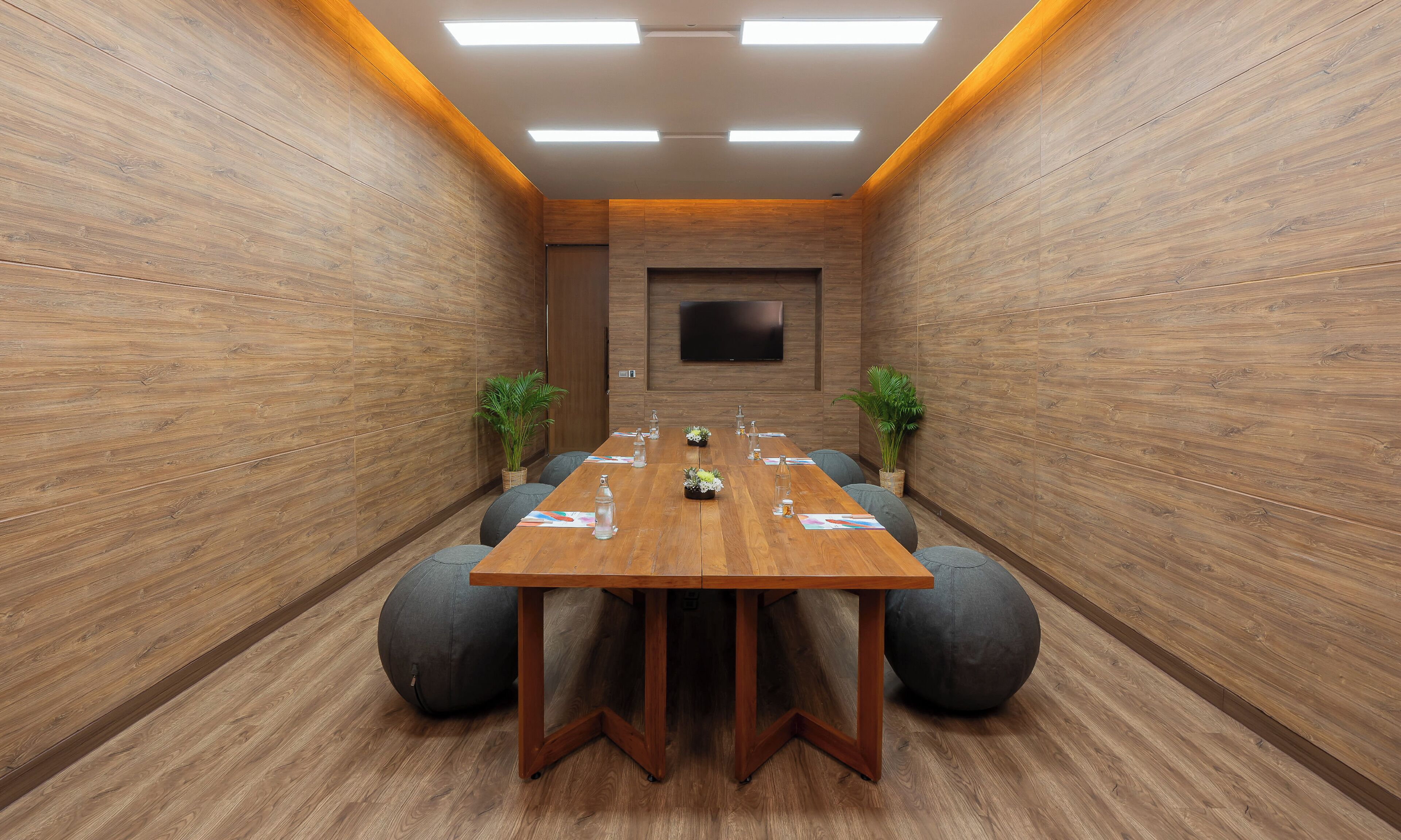 A conference room with a long wooden table, grey bean bag chairs, and potted plants.