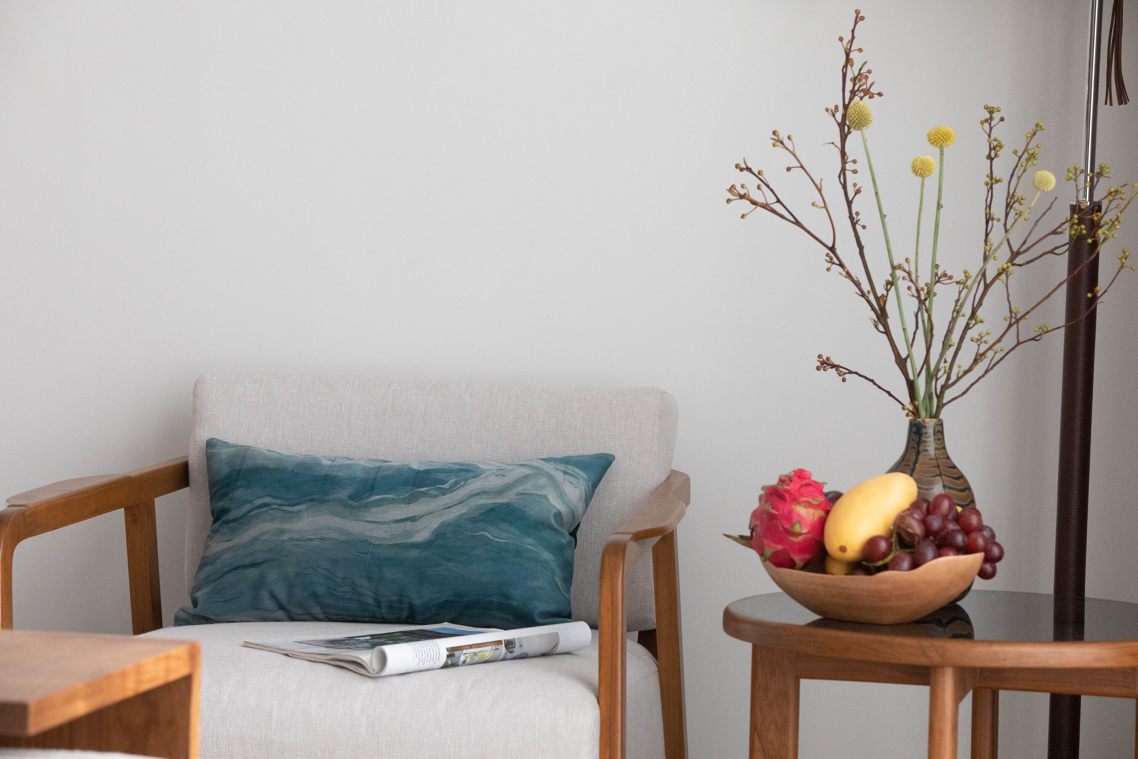 A room with a wooden chair, a cushion, a magazine, a vase with flowers, and a bowl of fruit.