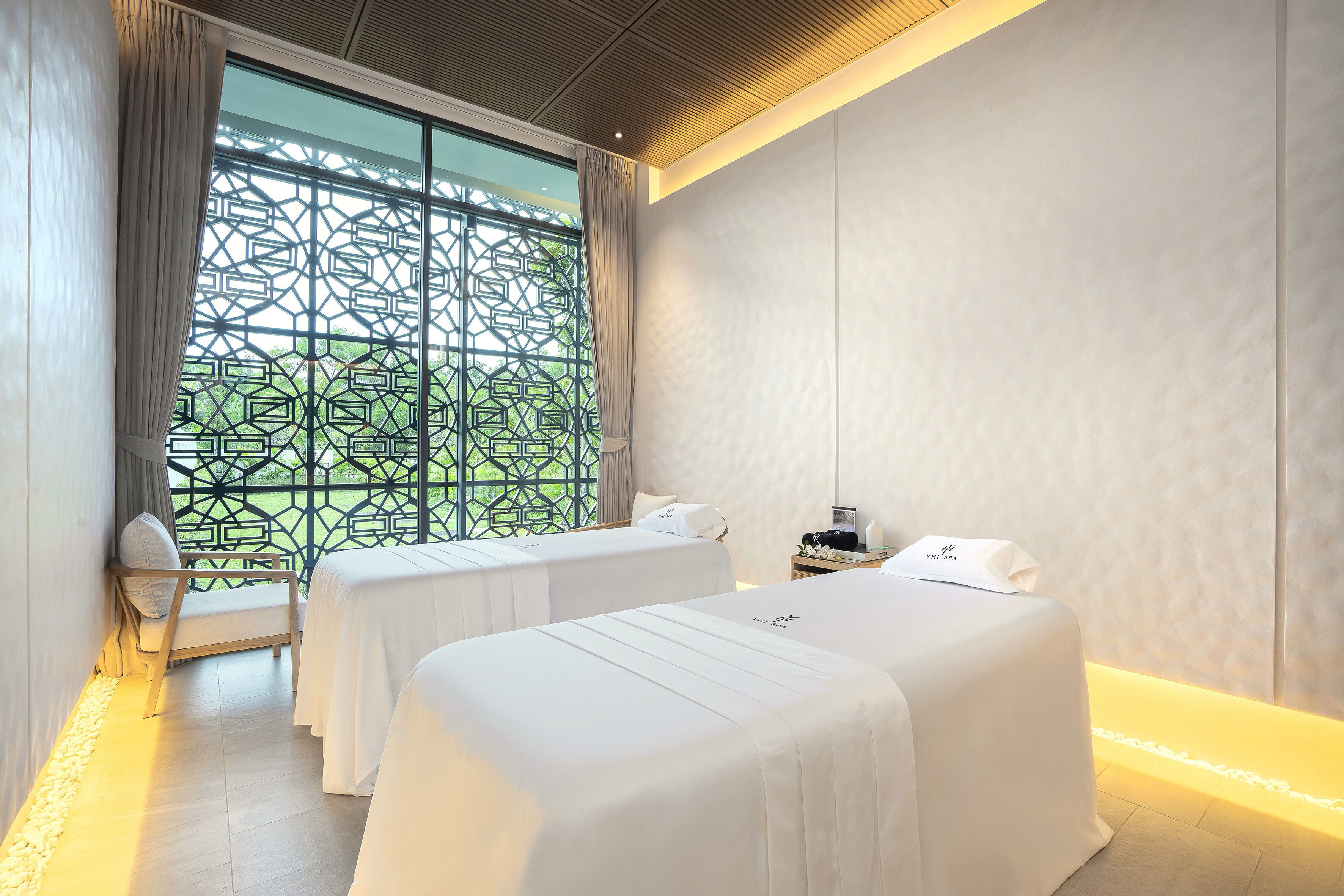 A hotel room with two beds, a large window with a geometric pattern, a bedside table with a lamp, and a wall-mounted TV.