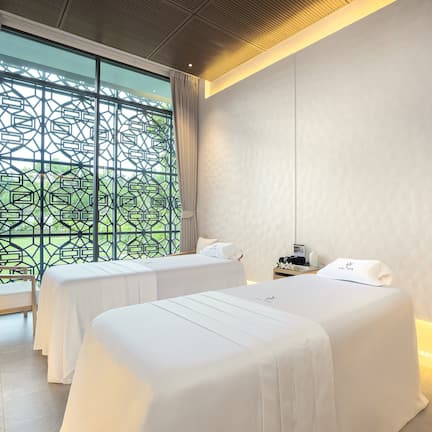 A hotel room with two beds, a large window with a geometric pattern, a bedside table with a lamp, and a wall-mounted TV.