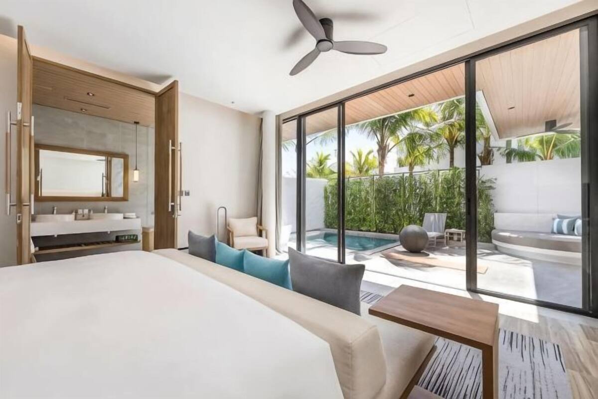 Quarto de hotel moderno com uma cama grande, banheiro com espelho, ventoinha no teto e vista para uma piscina e área de estar externa.