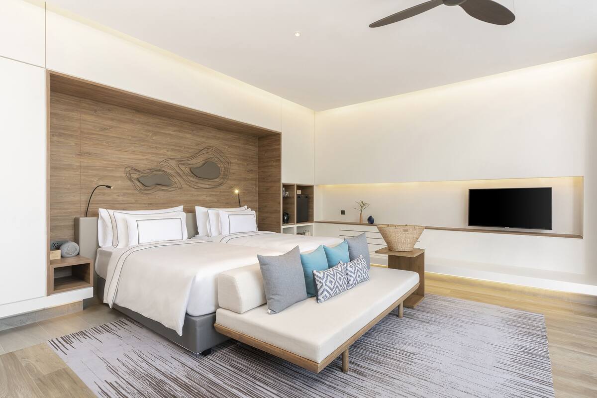 Quarto moderno com uma cama grande, mesas de cabeceira, um banco, uma TV e uma parede de destaque em madeira.